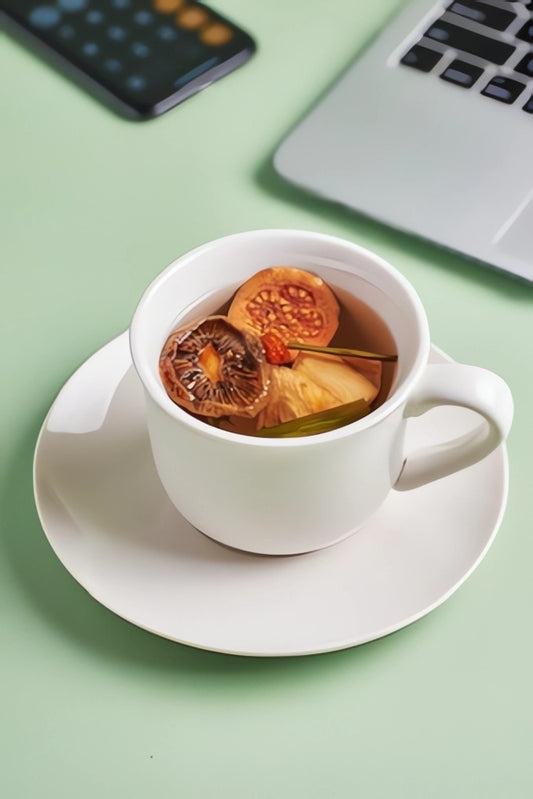"A white cup of infusion with slices of guava, kiwi, pineapple, and lemongrass floating inside. The cup sits on a pastel green background, next to a laptop and a calculator."