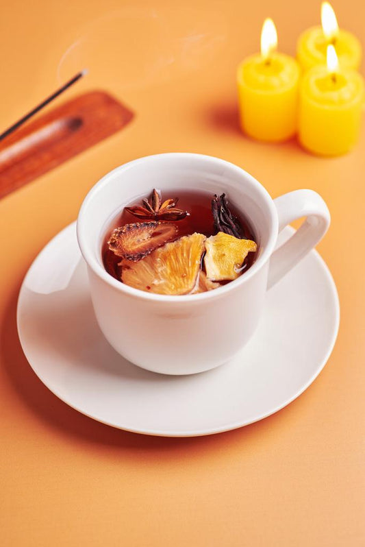 "A white cup of infusion with slices of pineapple, strawberry, star anise, and mango floating inside. The cup sits on a sunset orange background, next to three small, yellow lit candles and a wooden incense holder with a burning incense stick."